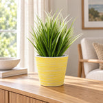 A potted artificial green grass plant in a yellow striped pot, placed on a wooden console table in a bright, modern living room.