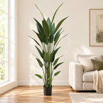 A tall artificial green plant with long, slender leaves in a black pot, styled in a bright living room next to a beige sofa.
