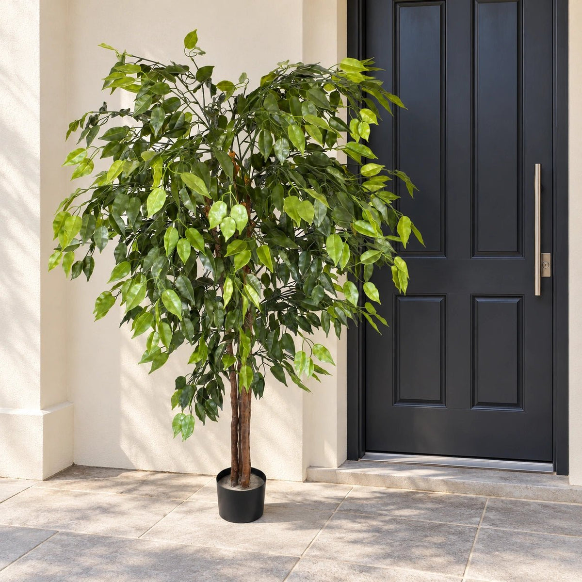 A potted artificial leafy tree with green foliage and a brown trunk, placed on a tiled porch next to a dark gray front door.
