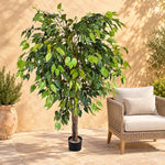 An artificial leafy tree in a black pot displayed on a patio next to a wicker chair, with a beige stucco wall background.