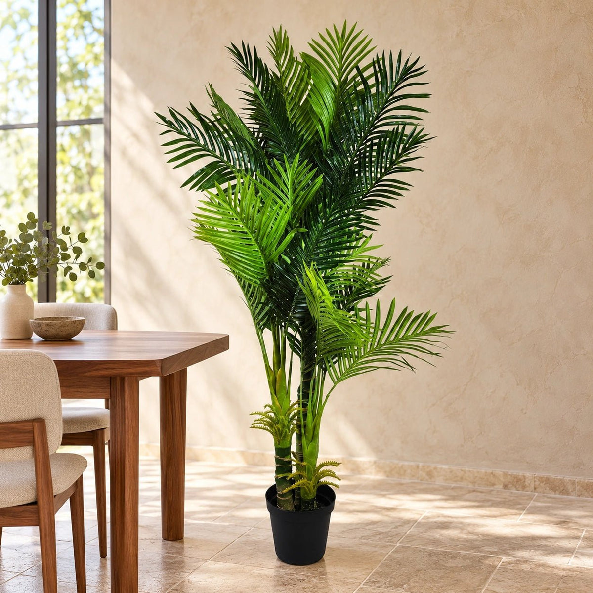 A tall artificial palm plant in a black pot placed beside a wooden dining table in a bright, modern room with beige walls and tiled floors.