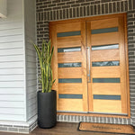 Modern double front doors with light oak wood panels and horizontal frosted glass inserts, set in a dark brick entryway with a potted snake plant and a welcome doormat.