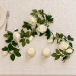 White rose garland with green leaves arranged on a cream tablecloth, styled with two small pillar candles and a partial plate and fork at the top left.