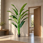 A tall artificial banana leaf plant in a black pot, styled in a modern, sunlit entryway with a wooden door open in the background.