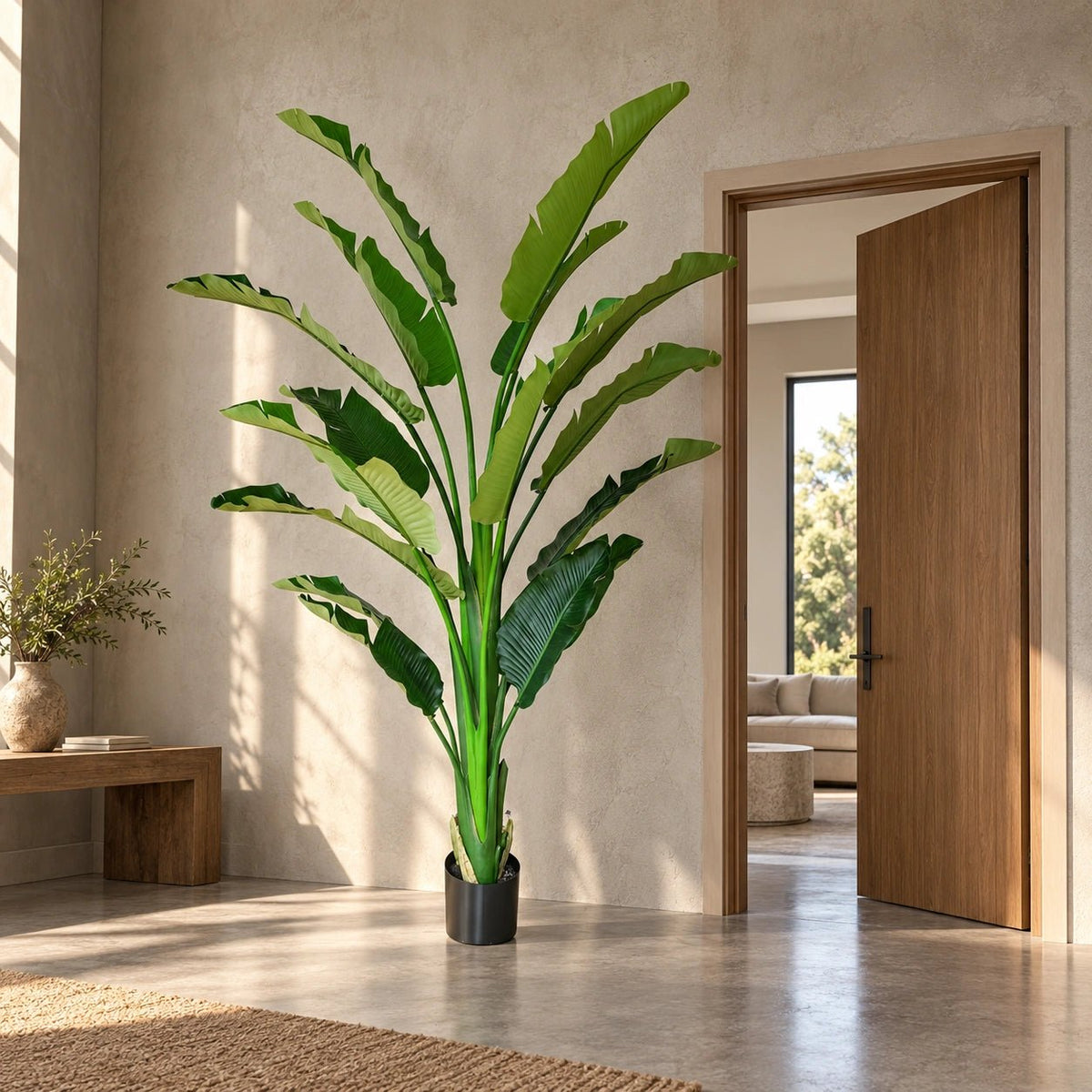 A tall artificial banana leaf plant in a black pot, styled in a modern, sunlit entryway with a wooden door open in the background.