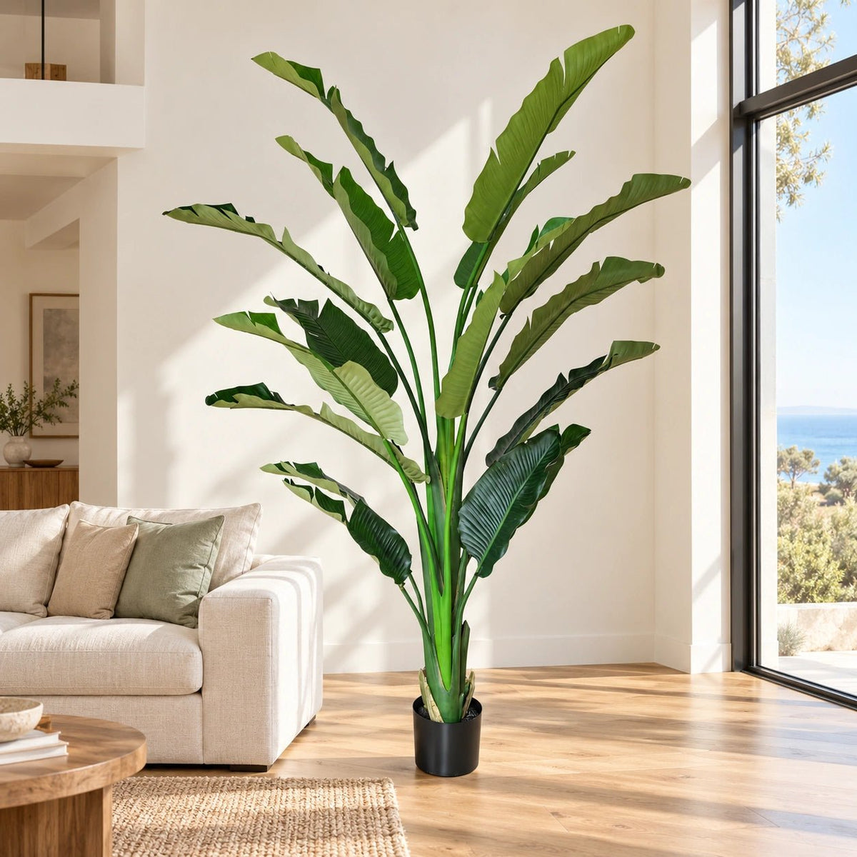 A tall artificial bird of paradise plant in a black pot, styled in a bright, modern living room next to a beige sofa and large windows.