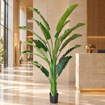 A large artificial banana leaf plant in a black pot, placed in a modern, sunlit hotel lobby with beige stone floors and a reception desk.