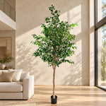 A tall artificial potted tree with green leaves and a light brown trunk, standing in a black pot on a wooden floor in a modern living room setting.