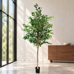 A tall potted artificial tree with green leaves and a light brown trunk stands in a modern room with large windows, a wood cabinet, and a light-colored floor.