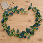 An artificial green leaf garland with textured dark and light green leaves, arranged in a circle on a light wood tabletop next to a small beige notebook.