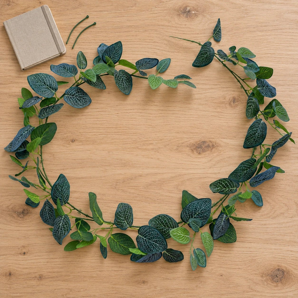 An artificial green leaf garland with textured dark and light green leaves, arranged in a circle on a light wood tabletop next to a small beige notebook.