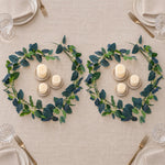 Two matching green and navy leaf garlands arranged on a beige tablecloth, each surrounding a small set of white pillar candles and matching candle holders, with plates, napkins, and gold-toned cutlery partially visible around the table.