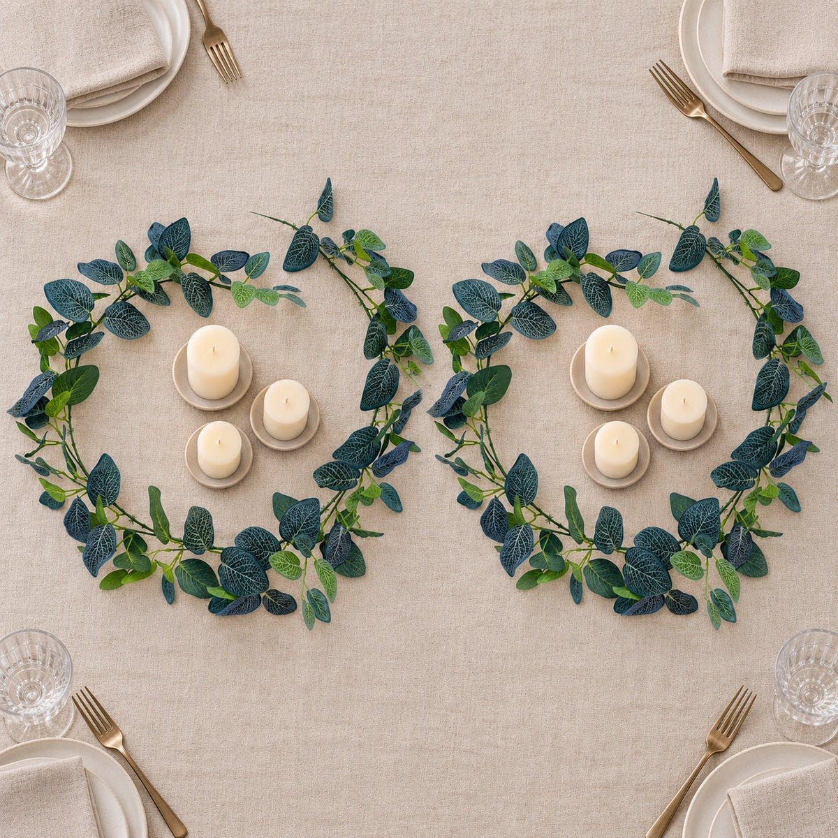 Two matching green and navy leaf garlands arranged on a beige tablecloth, each surrounding a small set of white pillar candles and matching candle holders, with plates, napkins, and gold-toned cutlery partially visible around the table.