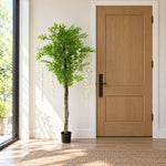 An artificial potted tree with green leaves and white wisteria-style hanging blooms stands in a bright entryway beside a light wood door and window.