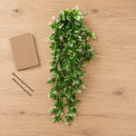 A green artificial leafy garland with white-tipped leaves is arranged on a light wood tabletop, with a small brown notebook and two pins placed to the left.