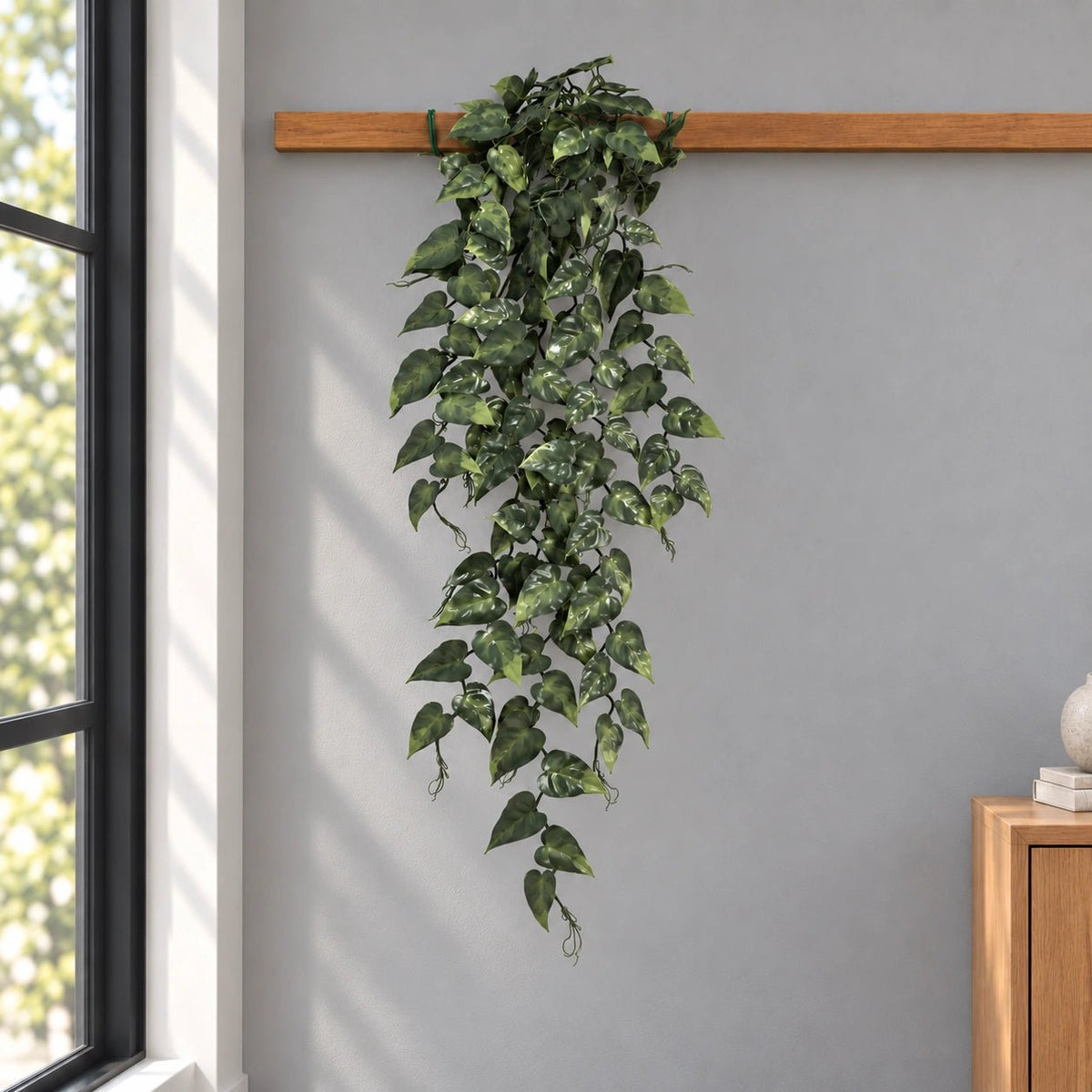 A long green trailing ivy vine with variegated light green leaves hanging from a wooden rod against a gray wall in a room setting.