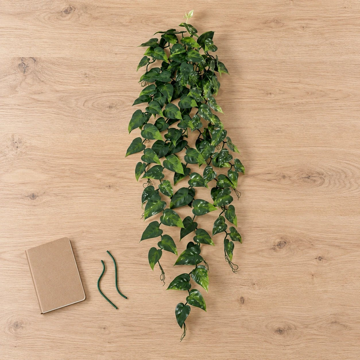An artificial trailing green ivy vine with variegated dark and light green leaves, displayed on a light wood grain surface next to a brown notebook and two green stems.