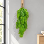 A long, lush green artificial palm frond hanging from a wooden peg on a light gray wall in a sunlit room, with a window and wooden furniture partially visible.