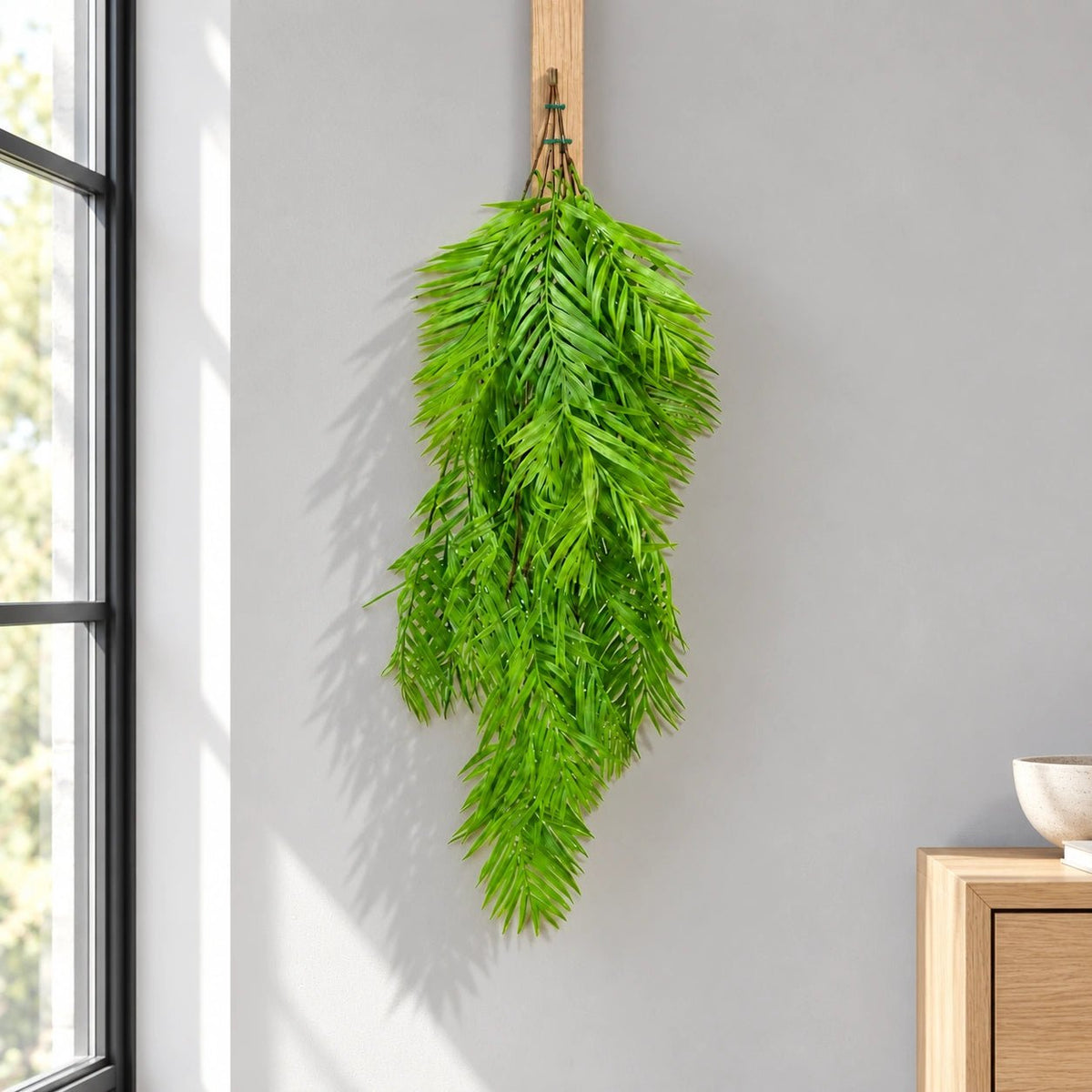 A long, lush green artificial palm frond hanging from a wooden peg on a light gray wall in a sunlit room, with a window and wooden furniture partially visible.