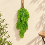 A green artificial palm frond hanging from a wooden hook on a textured beige wall, with soft shadows cast across the background.