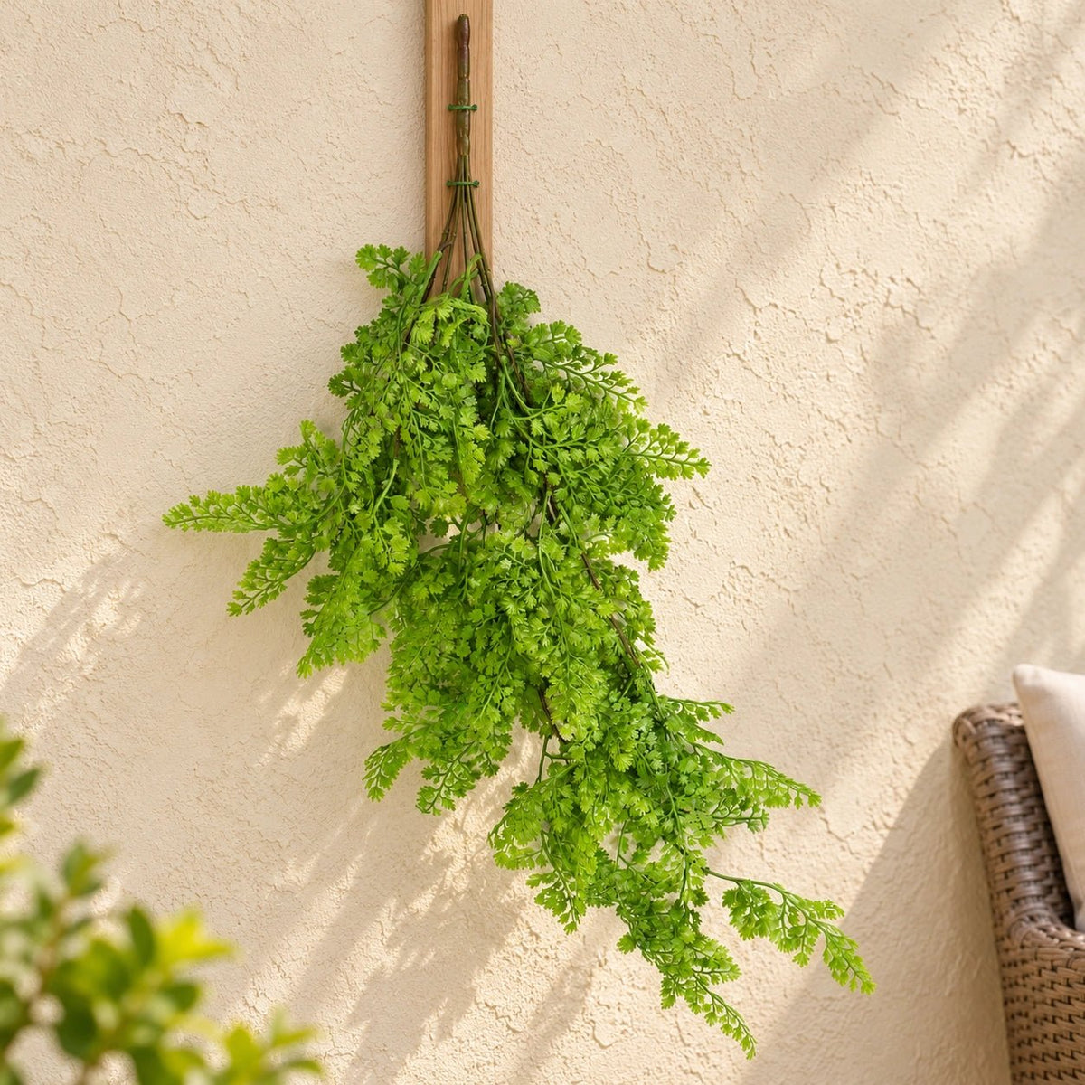A lush green artificial fern hanging from a wooden wall hook against a textured beige stucco wall, with soft shadows cast across the surface.