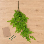 A bundle of artificial green fern branches laid flat on a light wood surface, with a small brown notebook and two green clips nearby as styling props.