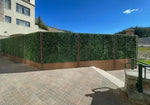 Green artificial hedge wall with wooden base against a building and clear blue sky