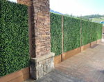 Artificial boxwood panels on a stone column and wooden fence with a scenic background.