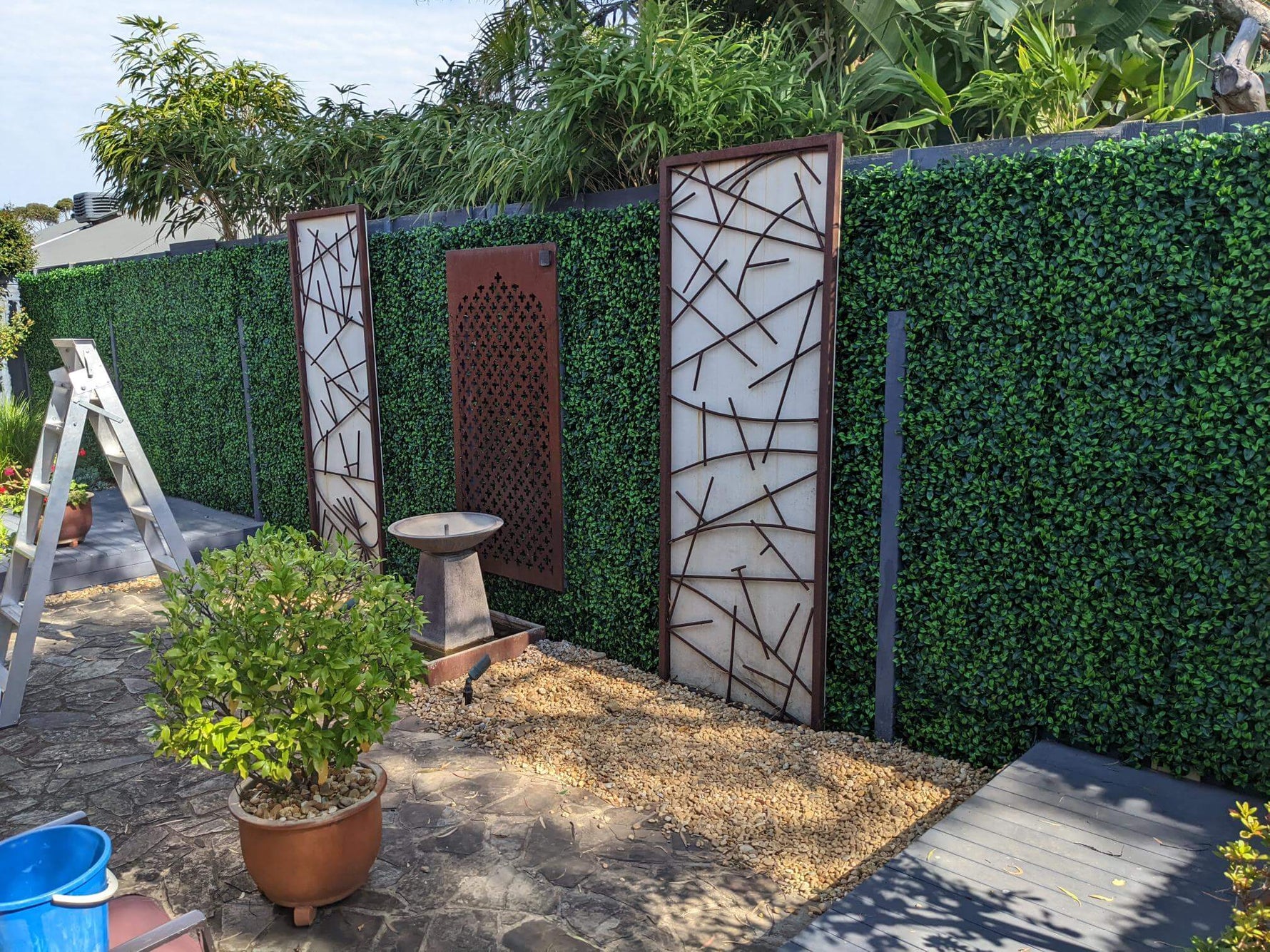 Decorative garden with geometric metal screens and potted plants against a green hedge.