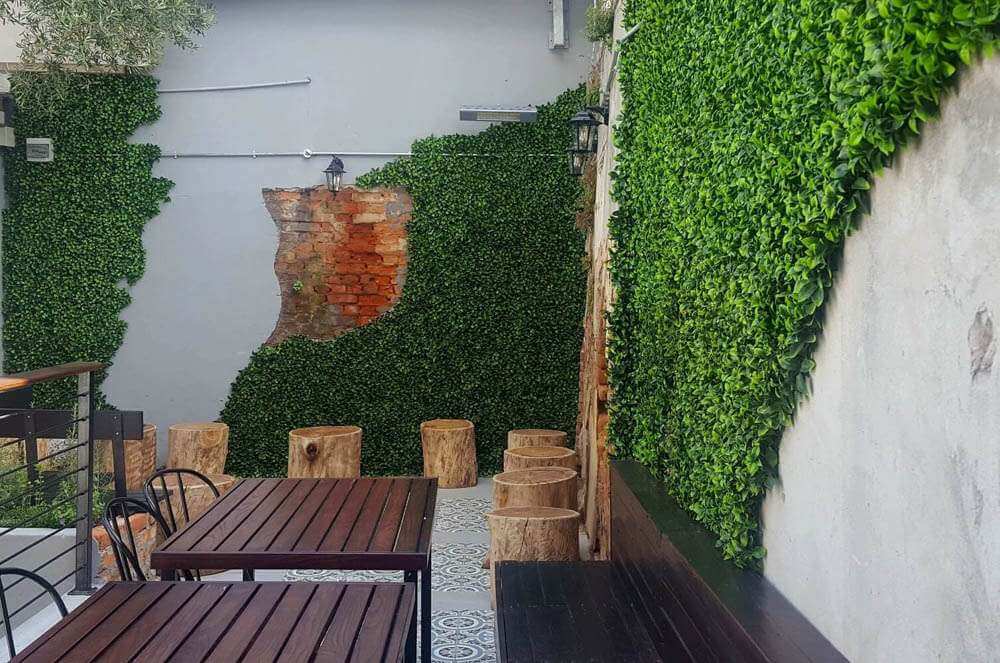 Outdoor seating area with wooden tables and chairs against a wall covered in greenery.