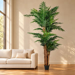 A tall artificial palm tree in a black pot, styled in a bright living room next to a beige sofa.