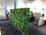 Green artificial plant wall in an office setting with people working.