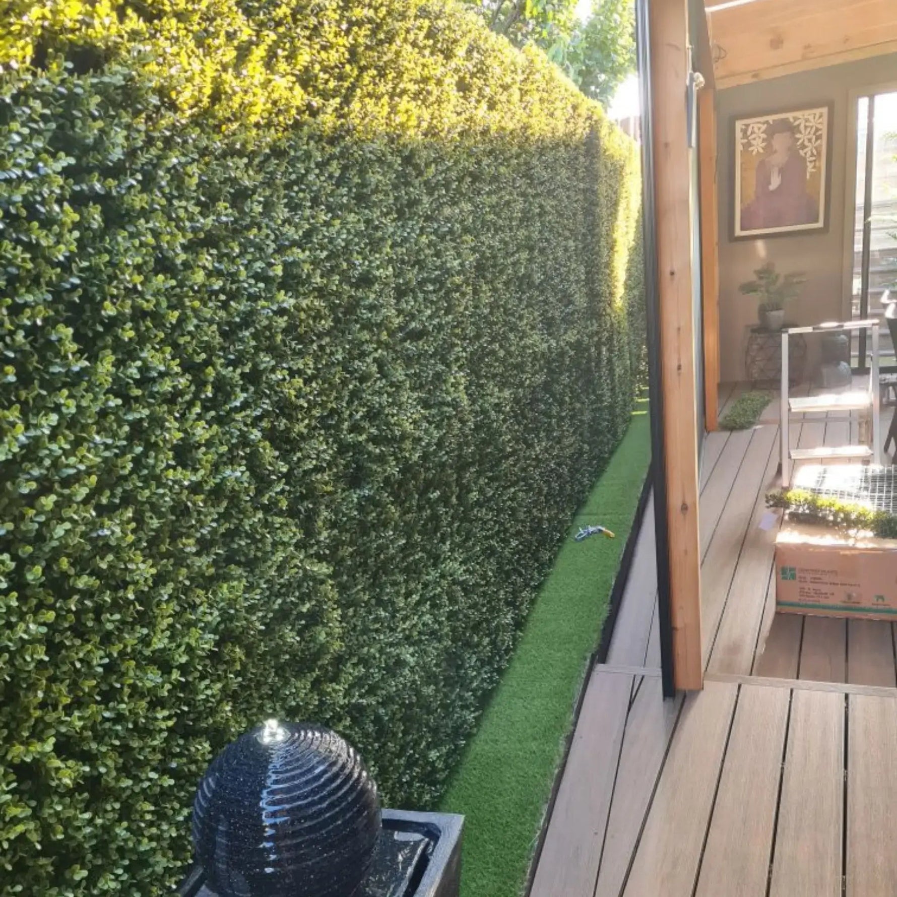 Artificial green hedge next to a wooden deck with a garden shed in the background