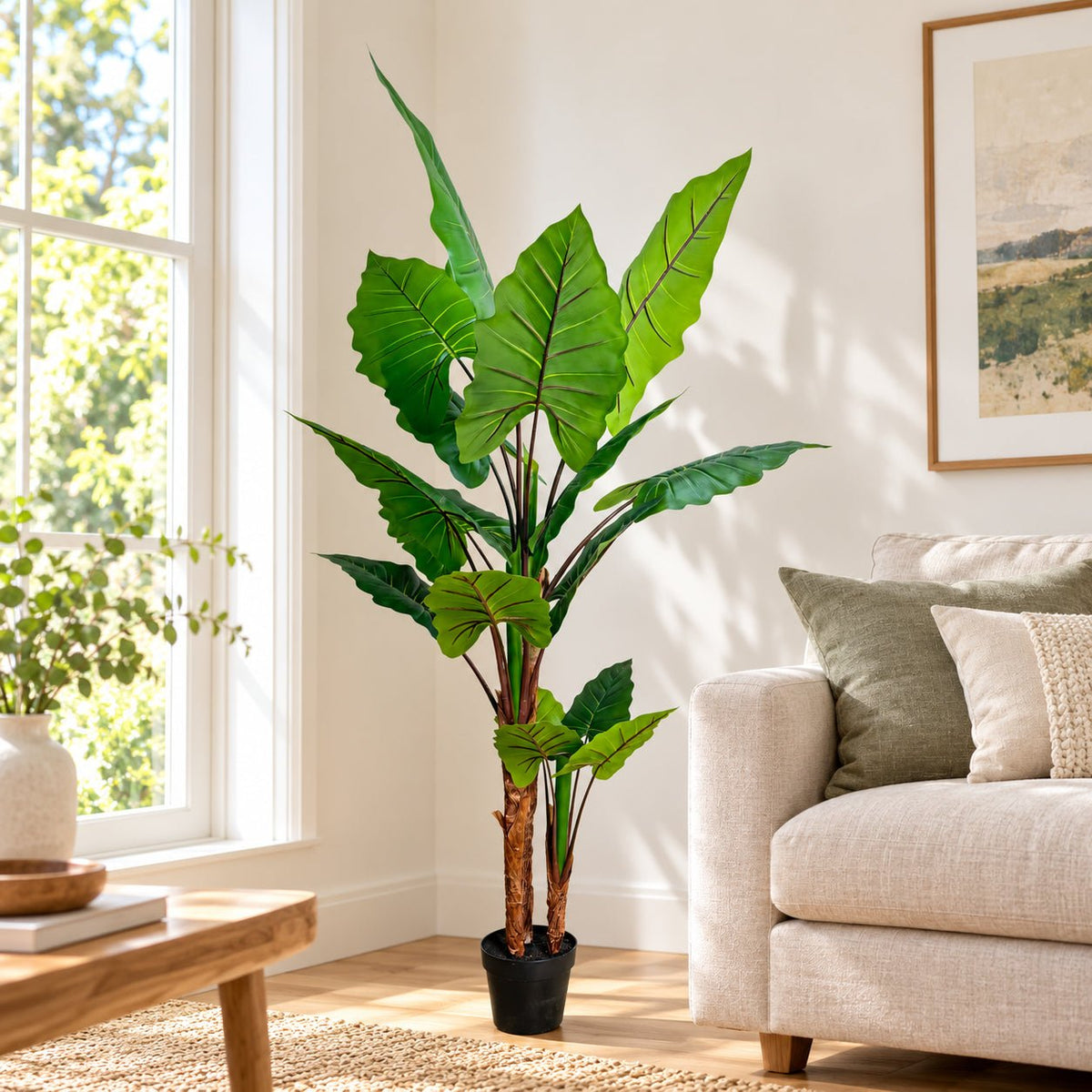 A tall artificial potted plant with large green tropical leaves and brown trunks, styled in a bright living room next to a beige sofa and a window.
