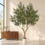 A large artificial olive tree with dense green foliage and a light brown trunk, potted in a white cylindrical planter, set in a minimalist room next to a wooden sideboard.