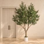 A potted artificial olive tree with dense green foliage and a brown trunk, displayed in a white cylindrical pot on a beige tiled floor in front of a cream-colored door and wall.