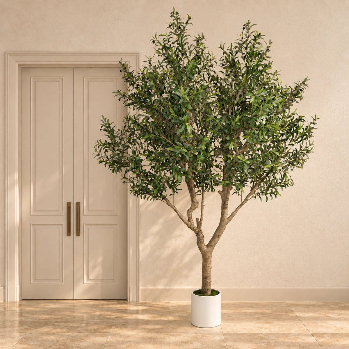 A potted artificial olive tree with dense green foliage and a brown trunk, displayed in a white cylindrical pot on a beige tiled floor in front of a cream-colored door and wall.