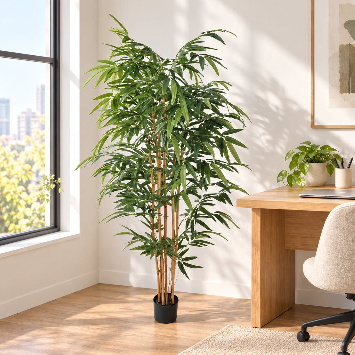 A tall artificial bamboo plant in a black pot stands in a bright home office next to a wooden desk and chair.