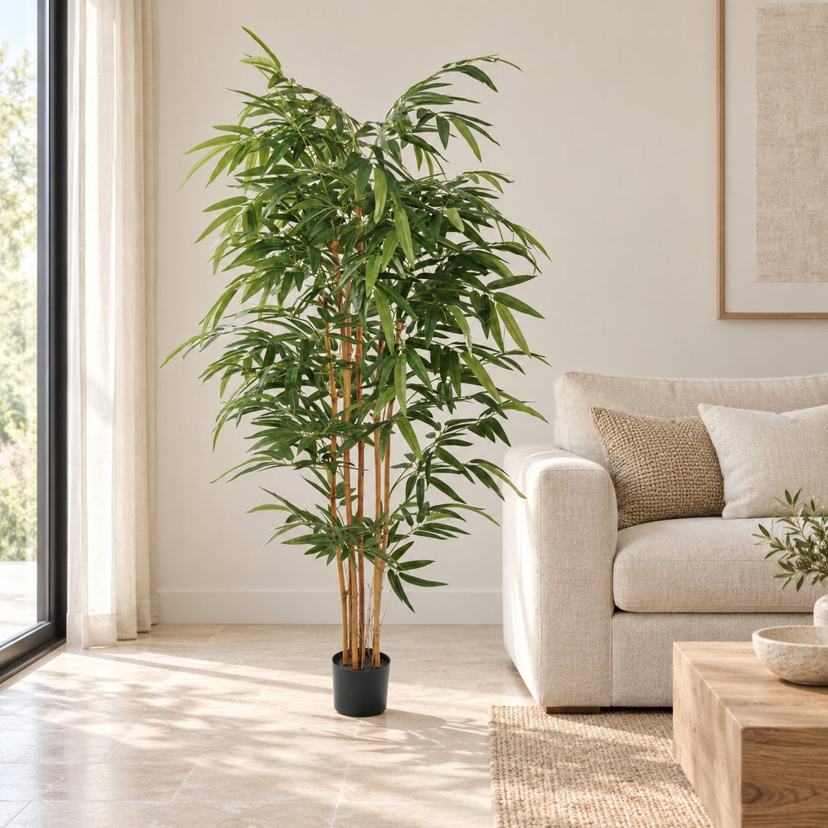 A tall artificial bamboo plant in a black pot, placed in a bright, modern living room next to a light beige sofa.