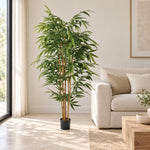 A tall artificial bamboo plant in a black pot, placed in a bright, modern living room next to a light beige sofa.