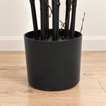 A matte black cylindrical planter holding several dark bamboo or decorative stems, placed on a light wood floor against a white wall.