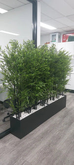 Artificial bamboo plant in a black planter box on a gray office floor.