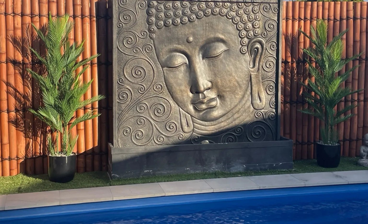 Decorative stone Buddha face wall fountain by a pool with plants on either side.