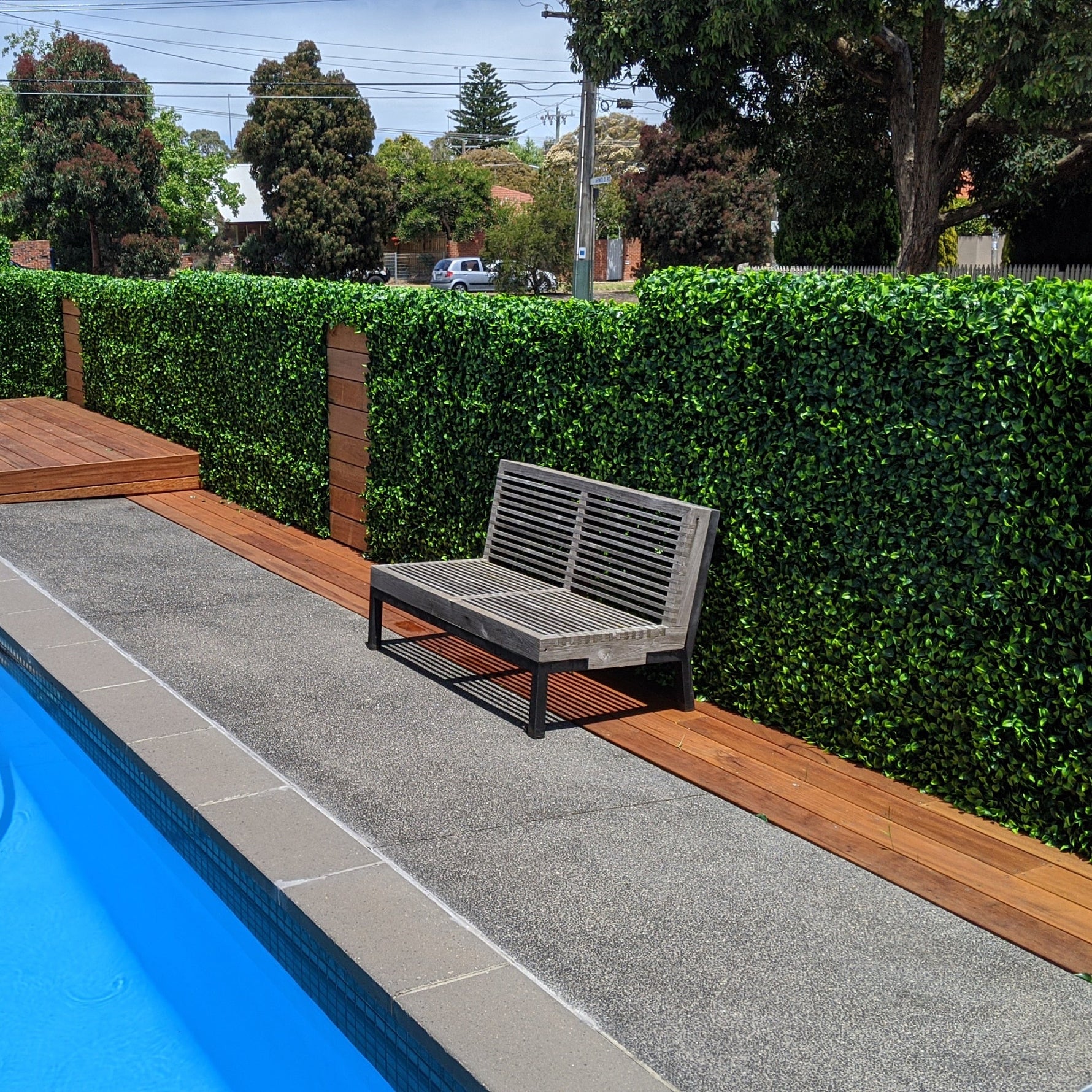 Gray outdoor bench by a pool with green hedges and trees in the background
