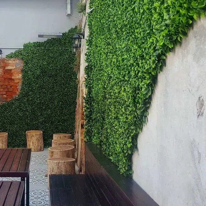 Outdoor seating area with wooden tables and stools against a green hedge wall.