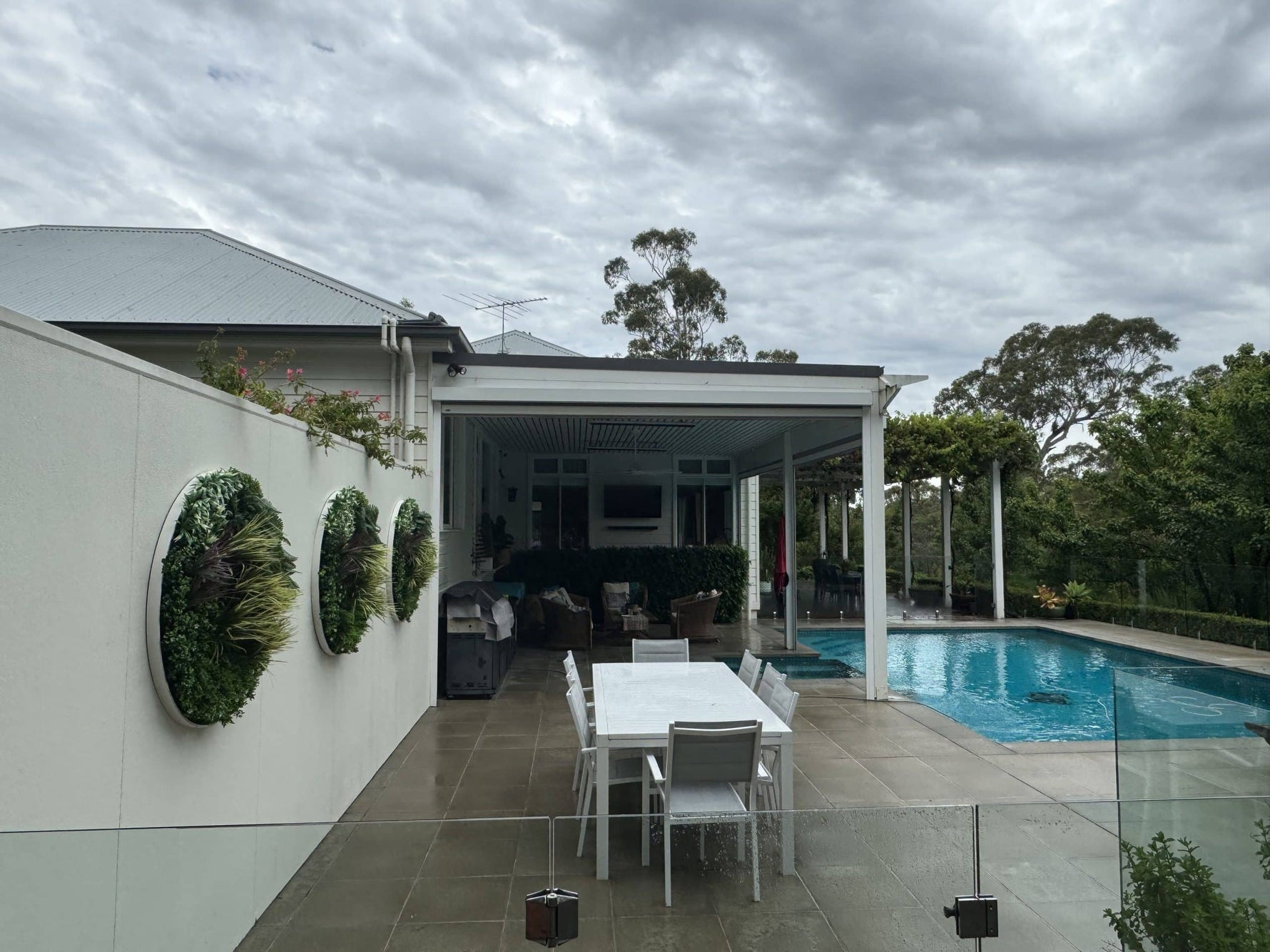 Outdoor patio area with a pool, table, chairs, and wall art on a cloudy day.