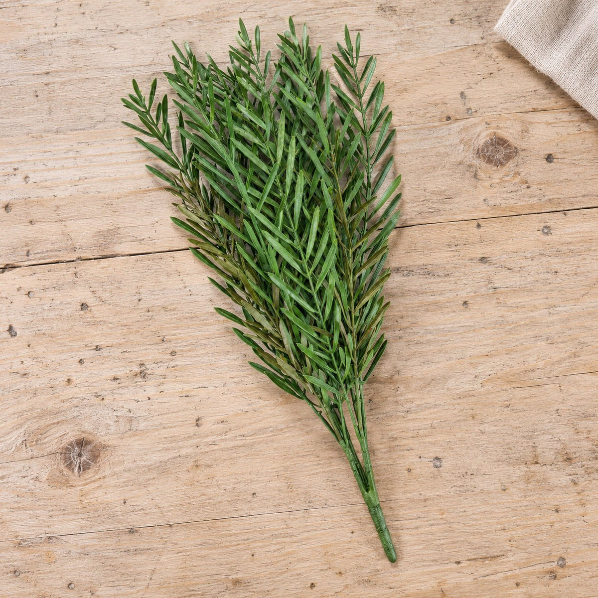 A bundle of artificial green pine branches with fine needle-like leaves, arranged on a light wood tabletop with a small beige fabric corner visible in the top right.
